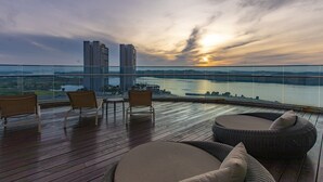 Un balcón con mobiliario de mimbre y vistas a un cuerpo de agua y edificios al atardecer.