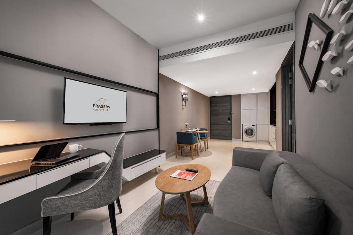 A modern hotel room with a flat-screen TV mounted on the wall, a desk with a laptop, a grey sofa, and a small round table with a magazine.