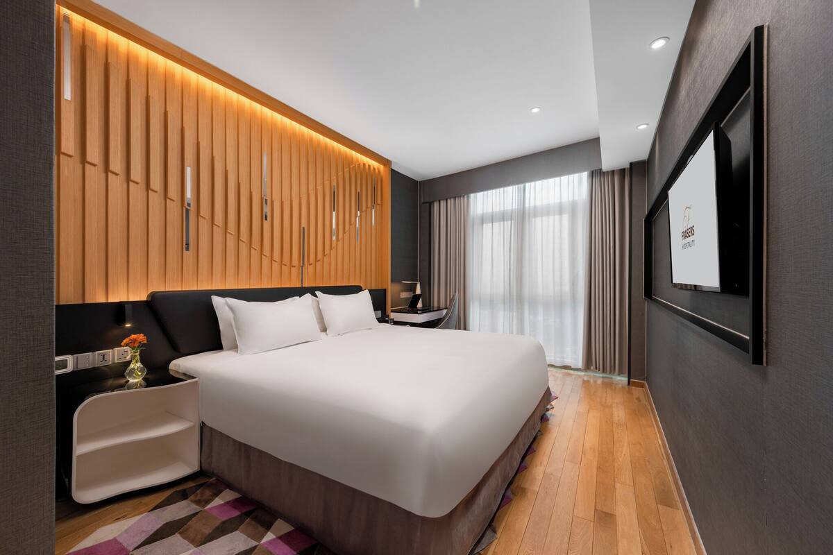 A modern hotel room with a large bed, a bedside table, a TV mounted on the wall, and a wooden panel design on the wall behind the bed.