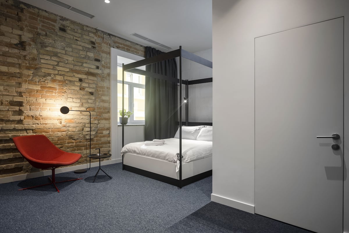 A modern hotel room with a brick wall, a red chair, a bed, and a door.