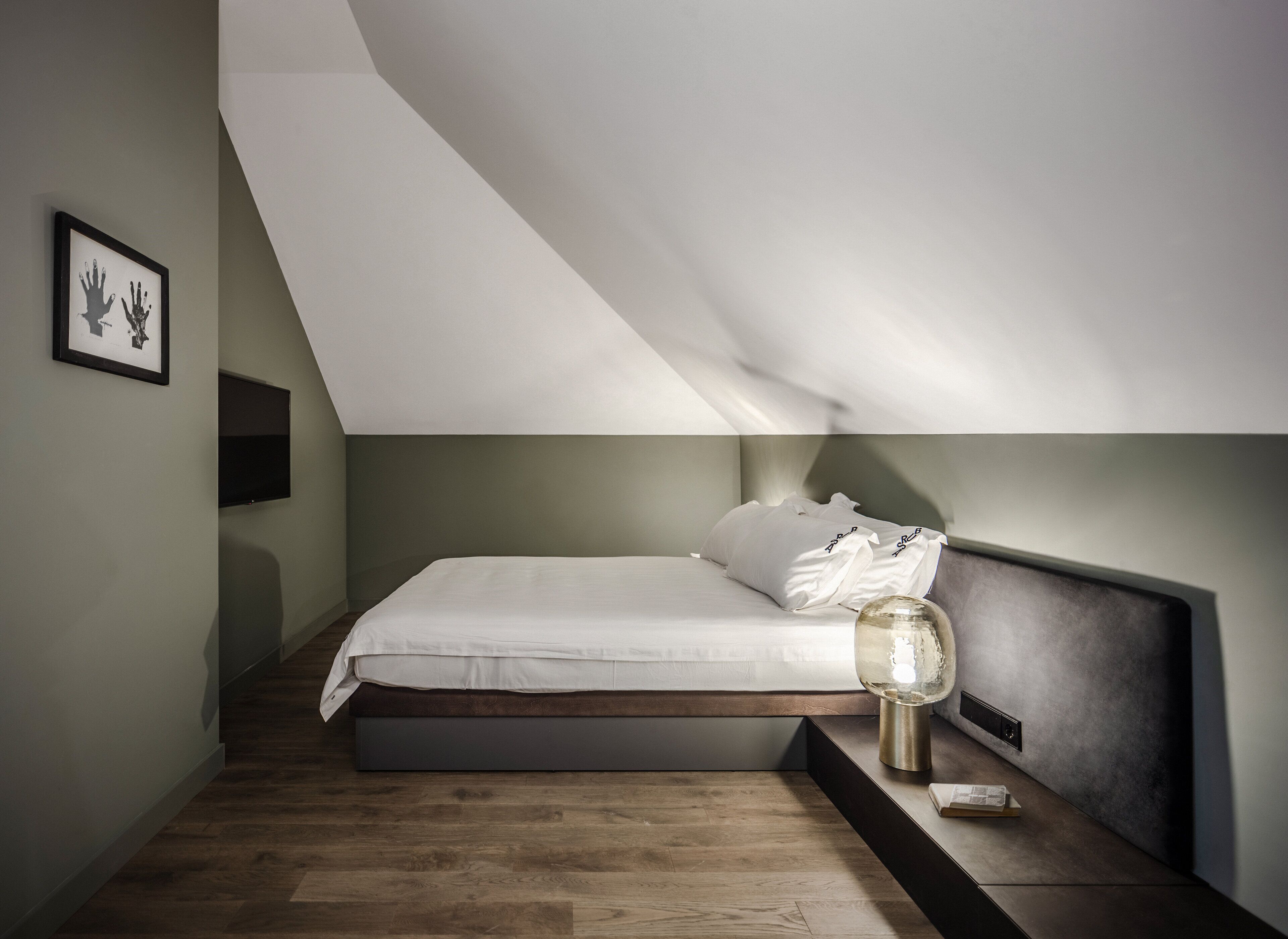 Un dormitorio moderno con techo inclinado, cama con ropa de cama blanca, mesita de noche con lámpara y un cuadro en la pared.