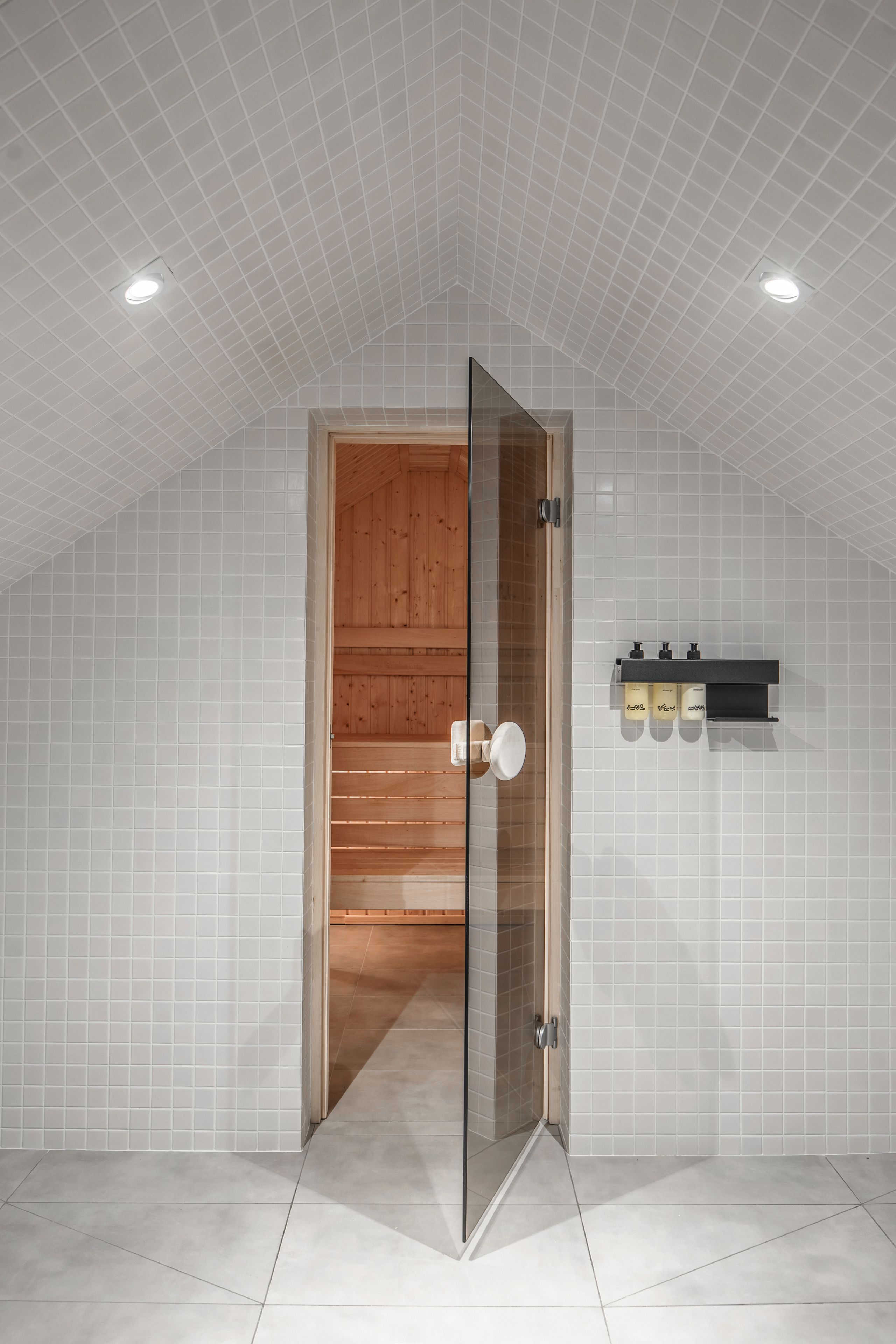 A modern bathroom with a glass door, white tiled walls, and a wooden panel wall. There are three bottles on a shelf.