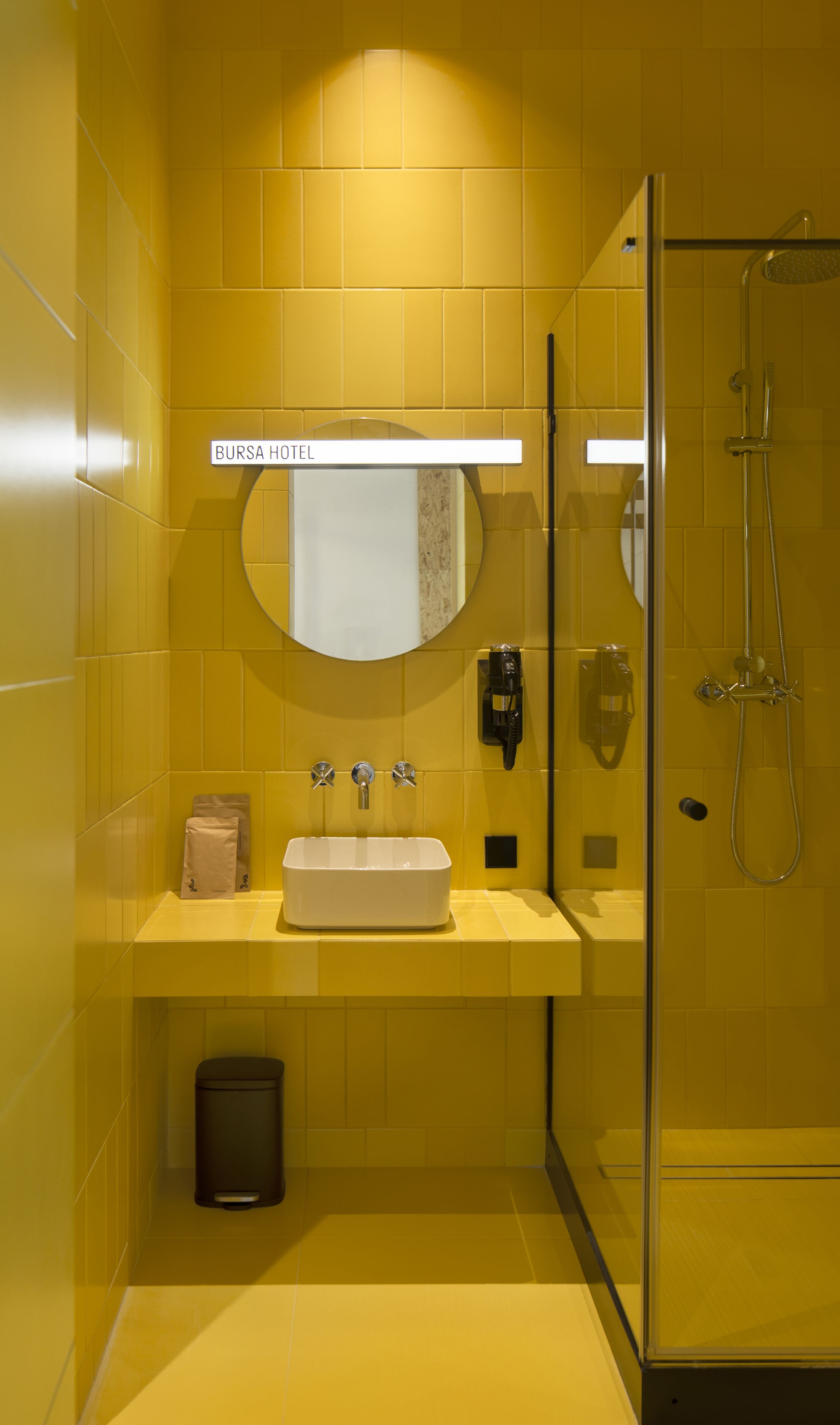 A bathroom with yellow tiles, a sink, a mirror, a glass shower enclosure, and a sign that reads "BURSA HOTEL".