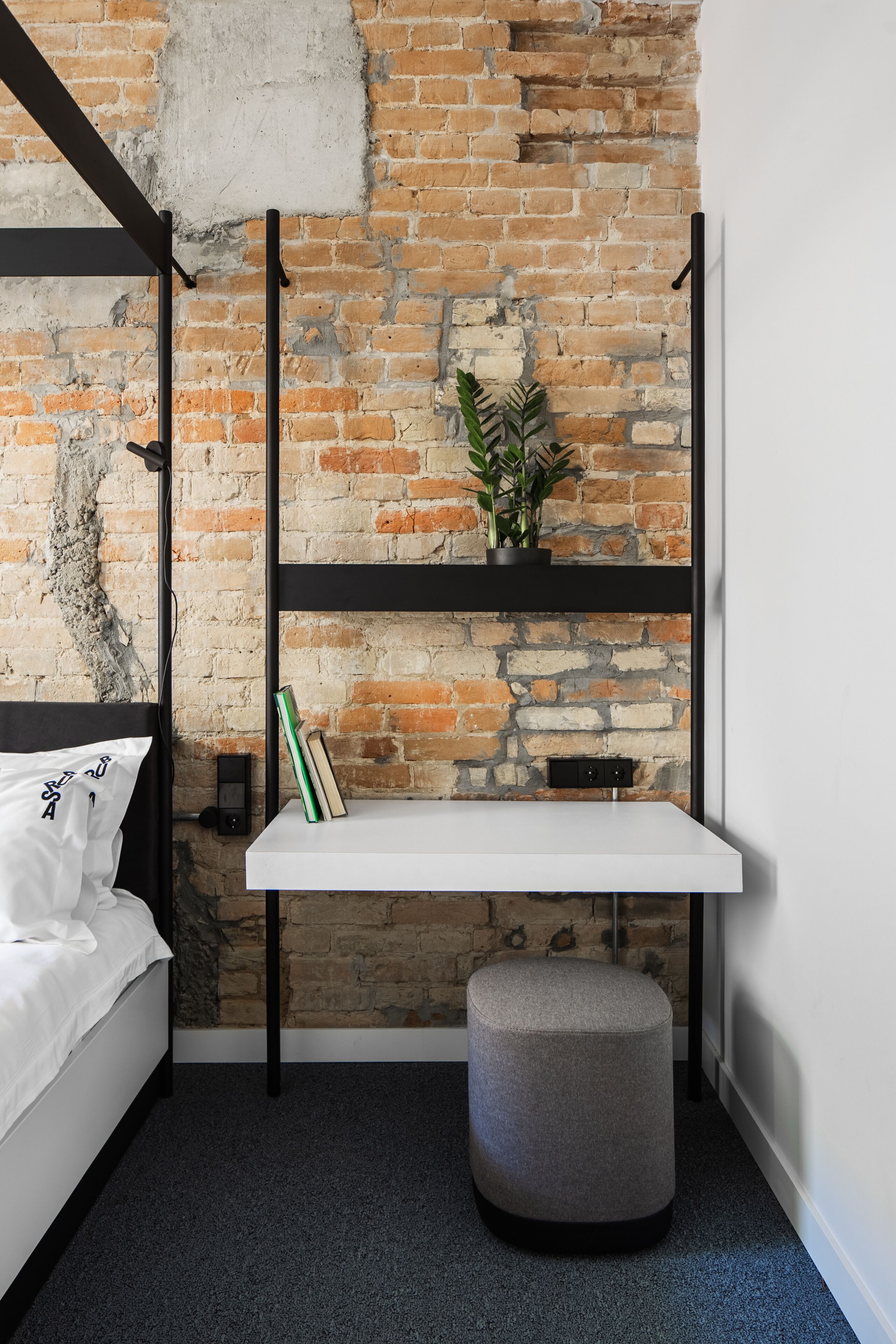 Ein Schlafzimmer mit Backsteinwand, einem Bett, einem weißen Regal mit Pflanze und einem grauen Pouf.