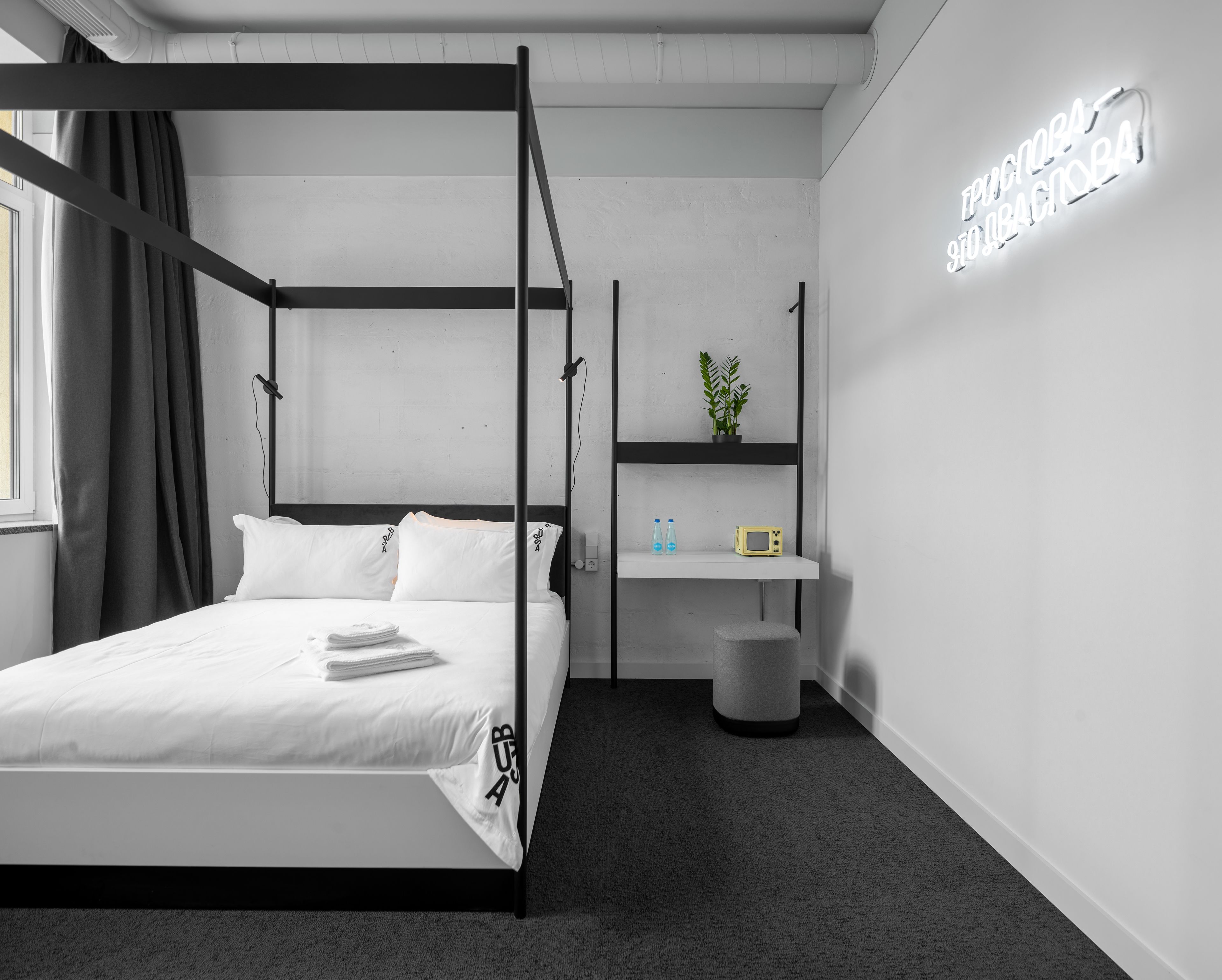 A modern bedroom with a four-poster bed, a small shelf with toiletries, and a neon sign on the wall.