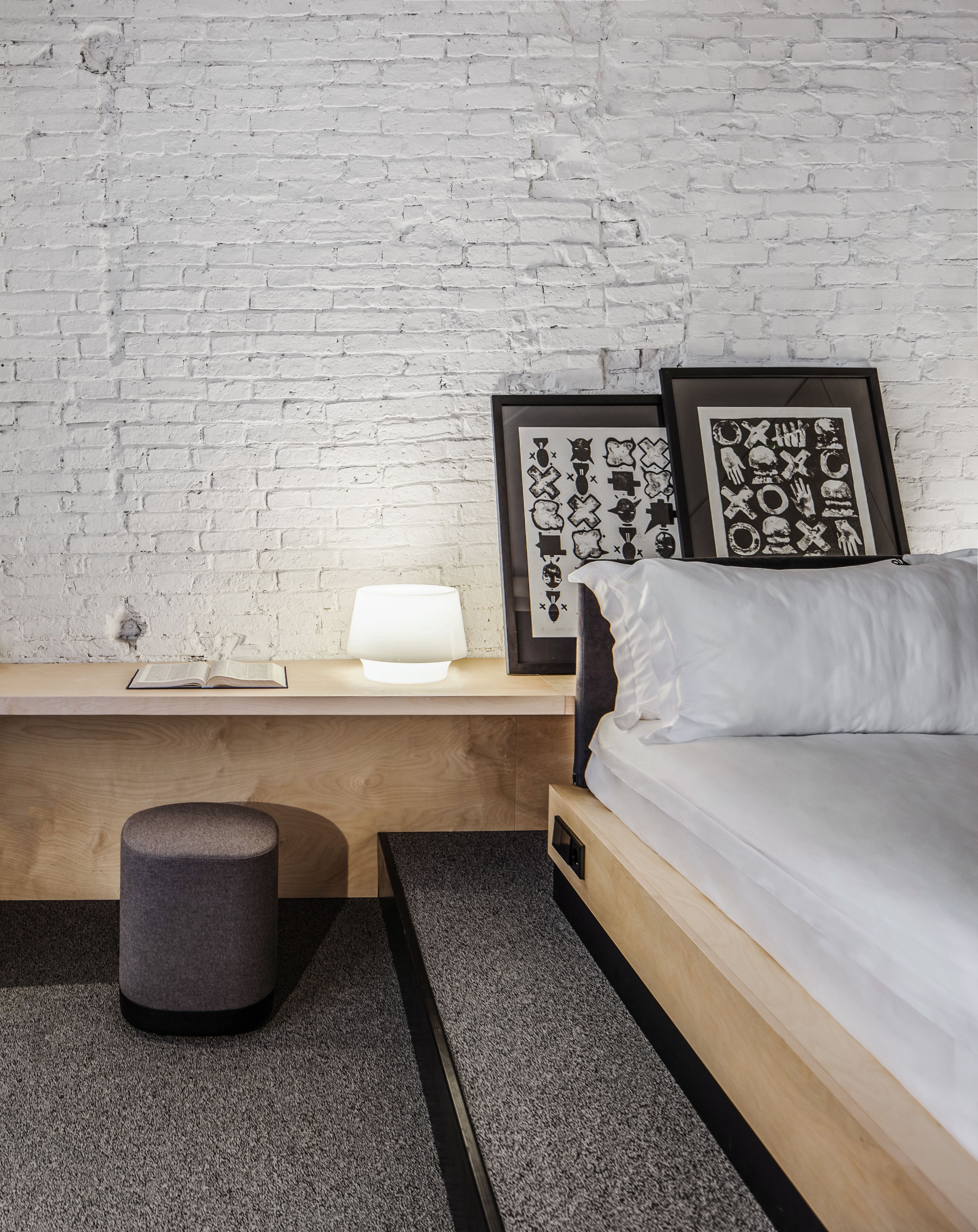 A modern bedroom with a white brick wall, a wooden nightstand with a lamp, a framed picture, a grey ottoman, and a bed with white bedding.