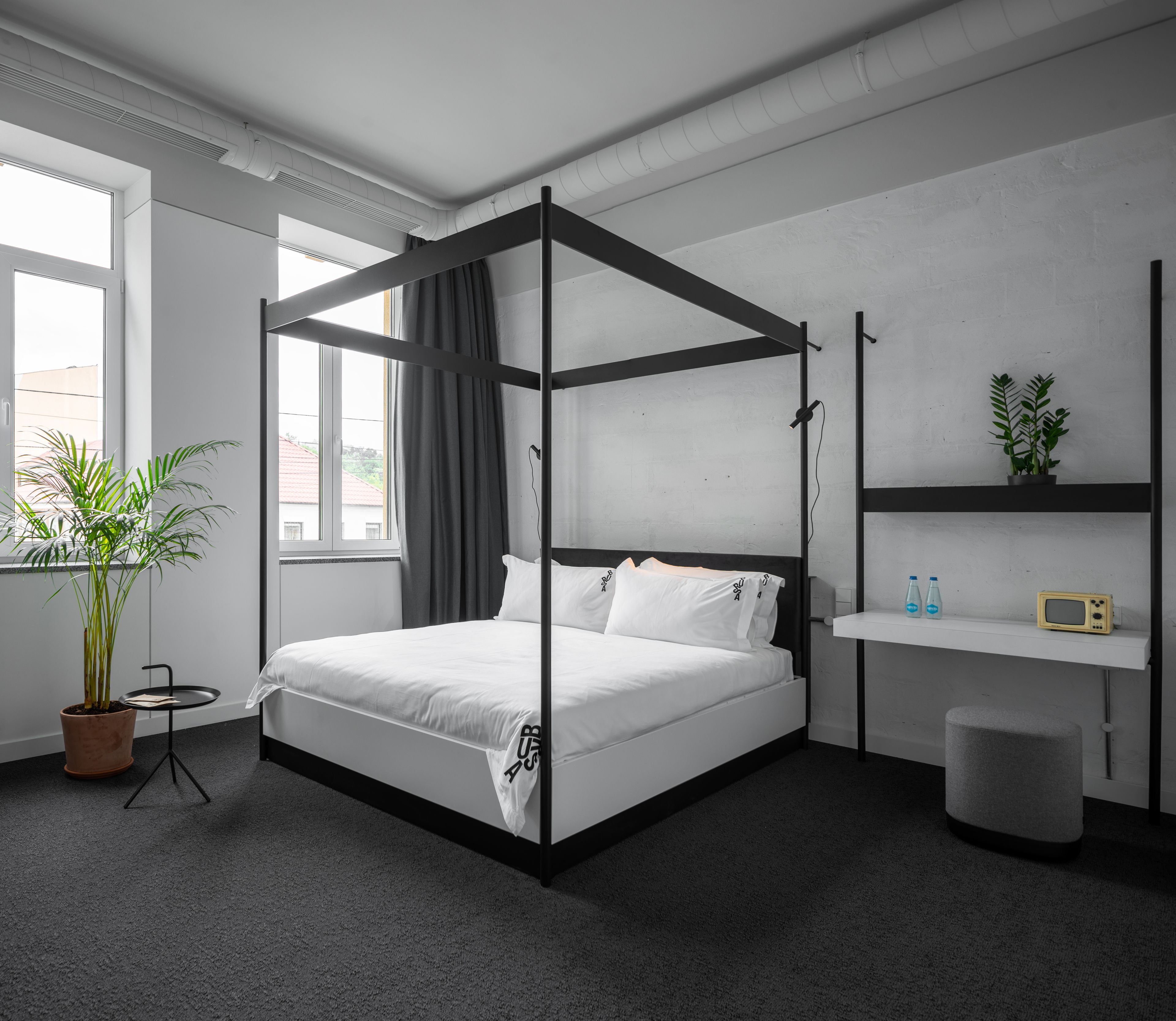 A modern bedroom with a four-poster bed, a small round table, a potted plant, a shelf with bottles and a radio, and a window with curtains.
