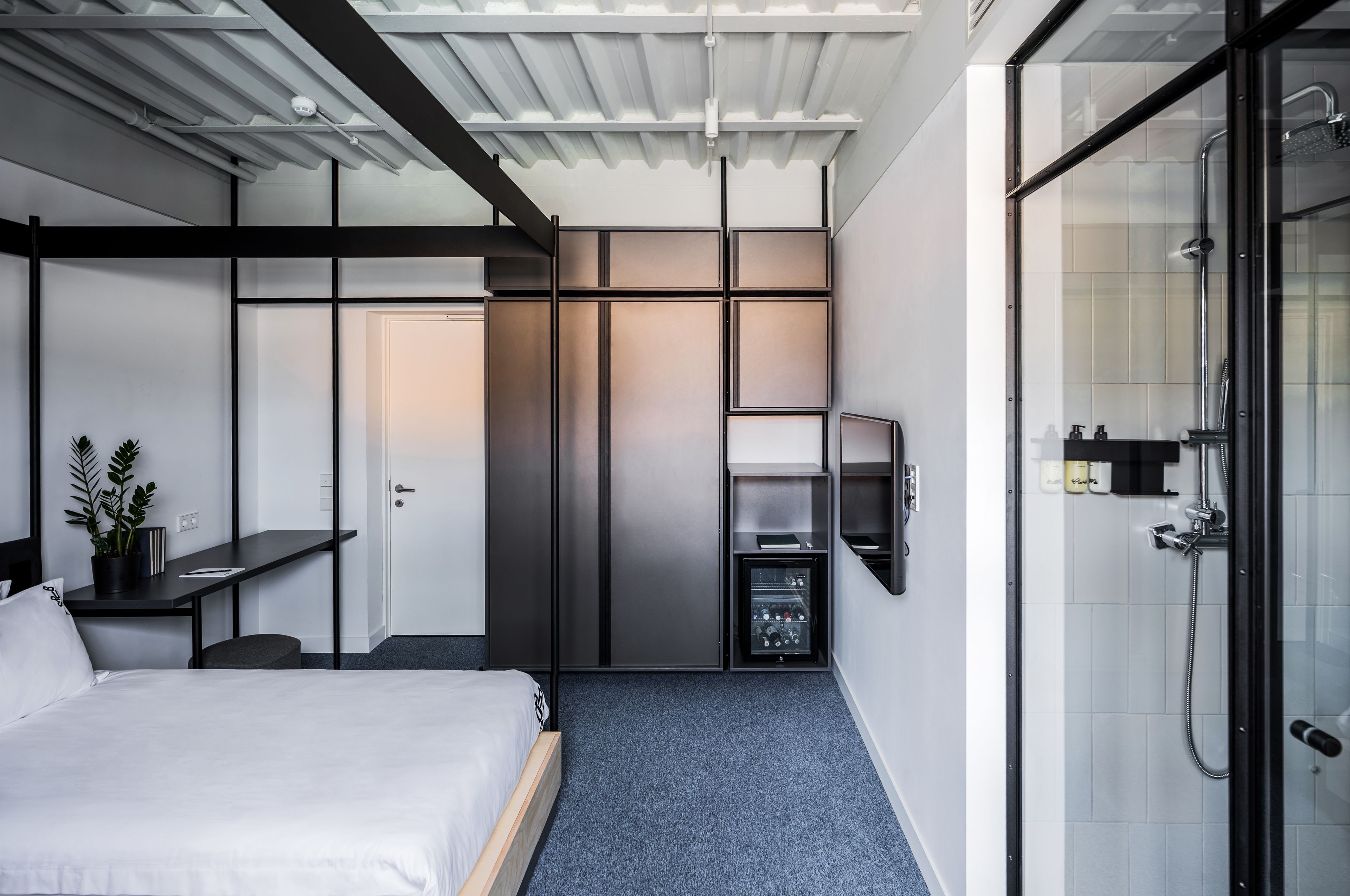 A modern hotel room with a bed, a desk, a wardrobe, and a glass shower area.