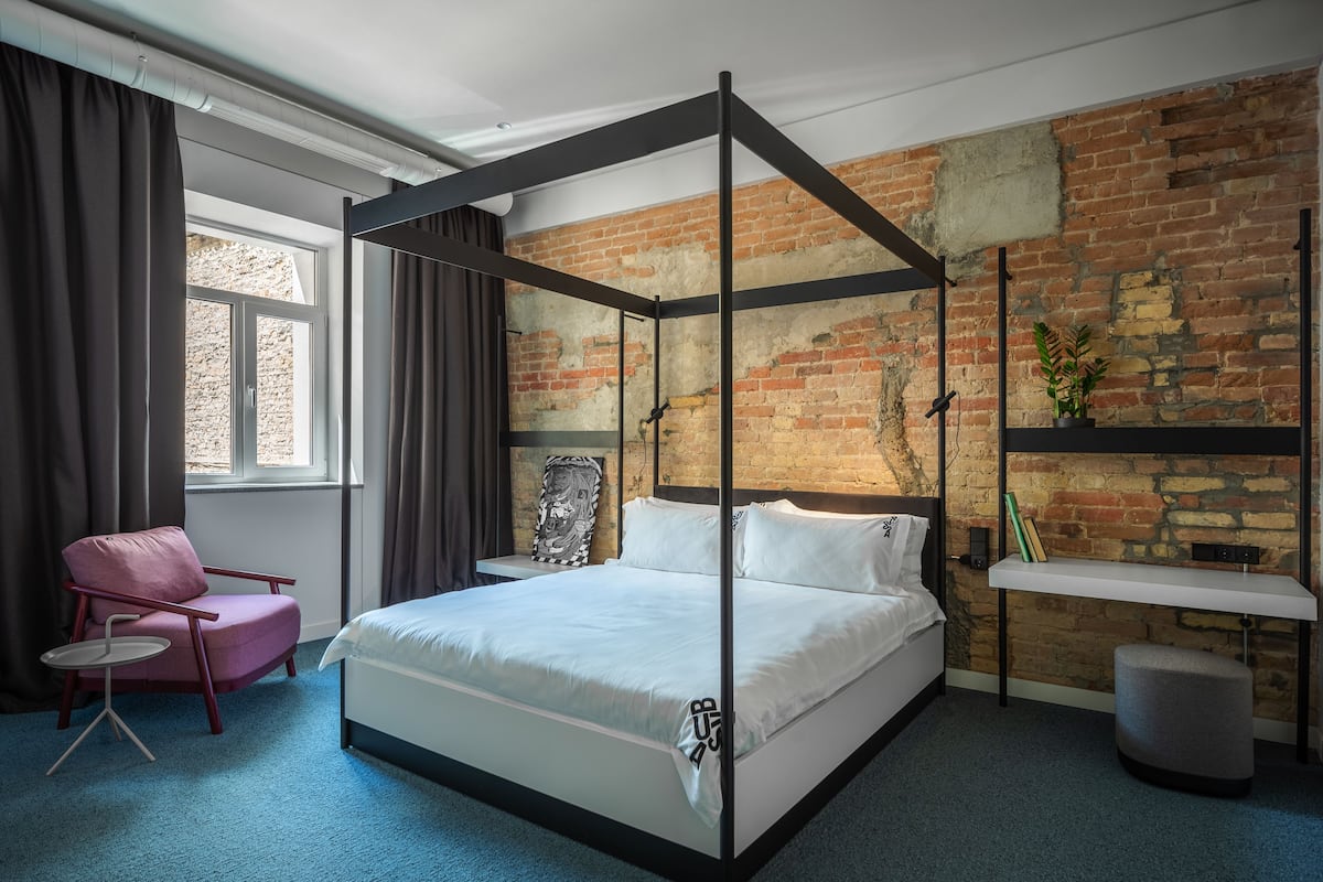 A modern bedroom with a four-poster bed, a pink chair, a small table, and a brick wall.