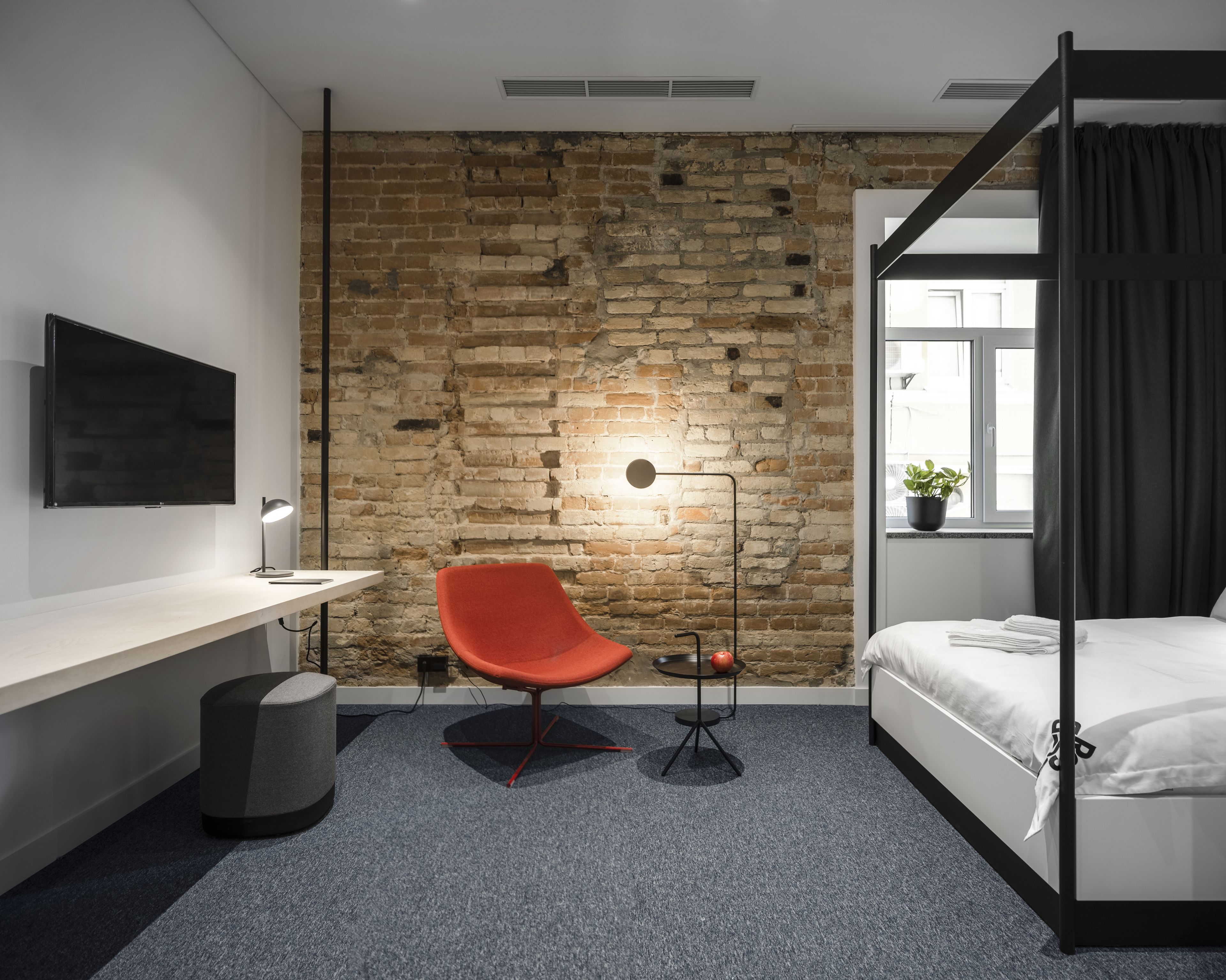 A modern hotel room with a brick wall, a desk with a chair, a bed, and a window with curtains.