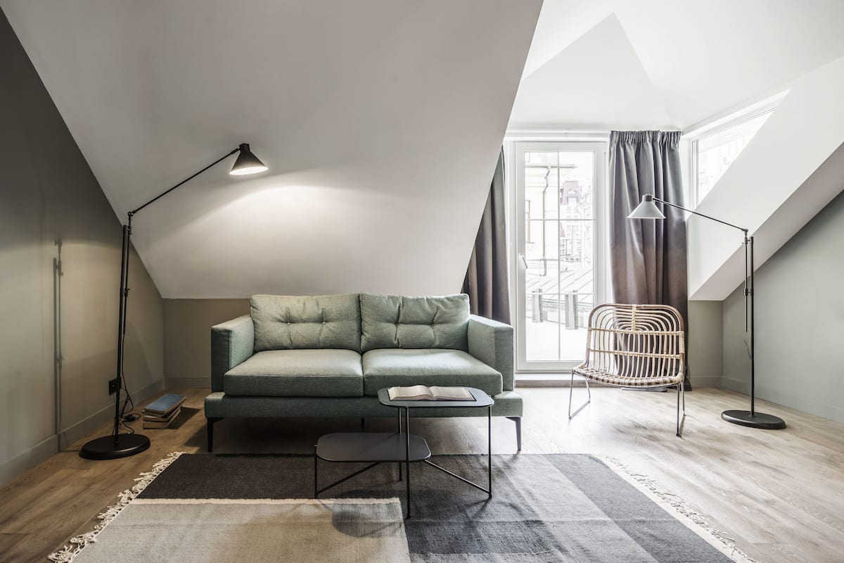 A modern loft bedroom with a green sofa, a small side table, a floor lamp, a chair, and a large window with curtains.