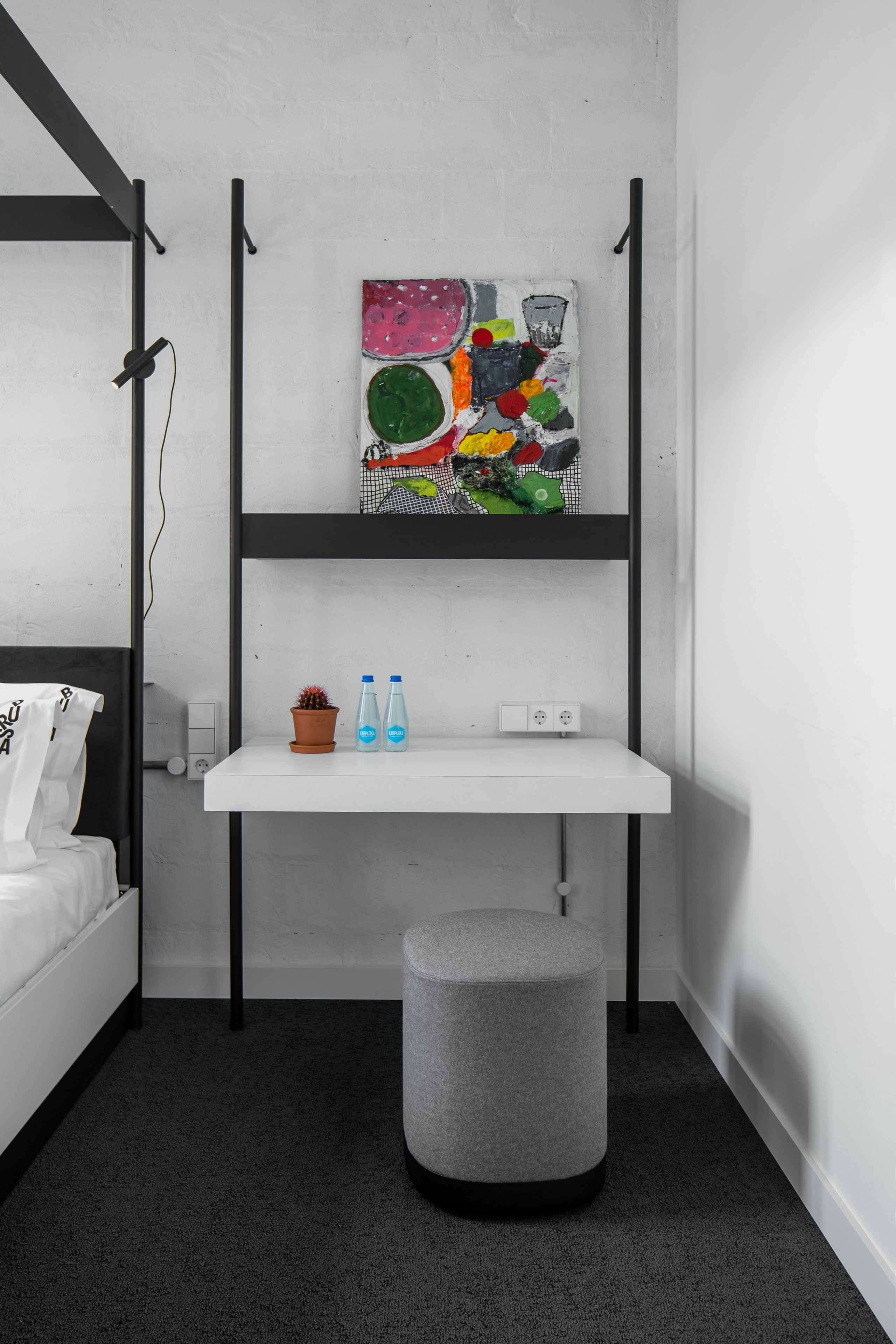 Una stanza moderna e accogliente con un letto a castello, una scrivania bianca con due bottiglie, un quadro astratto dai colori vivaci e un pouf grigio.
