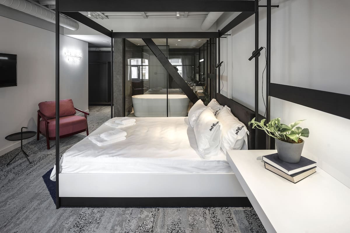 A modern hotel room with a large bed, a small seating area, a bathtub, and a potted plant.