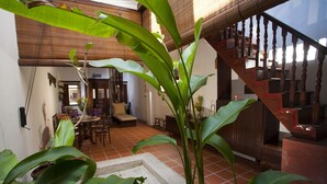 Un salón amplio con suelo de baldosas, una escalera de madera y grandes plantas verdes.