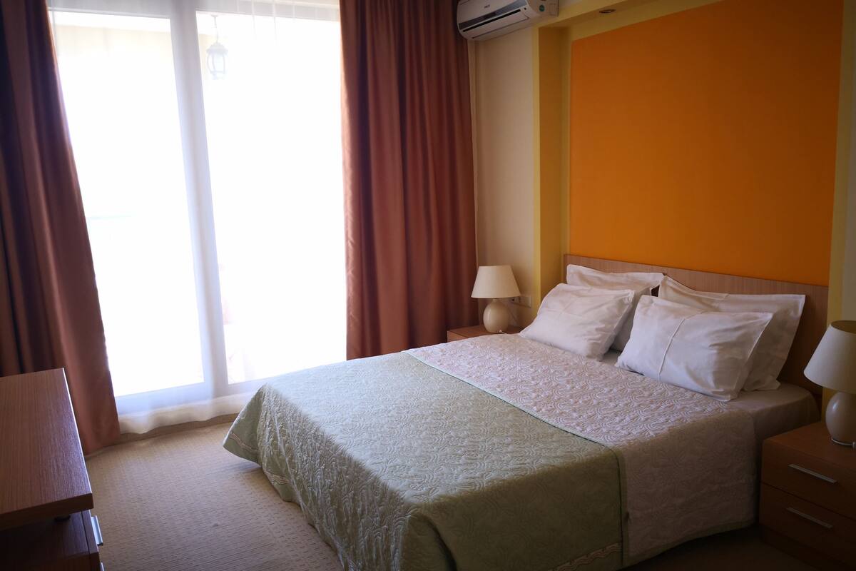 A hotel room with a bed, a nightstand, a lamp, and a window with curtains.