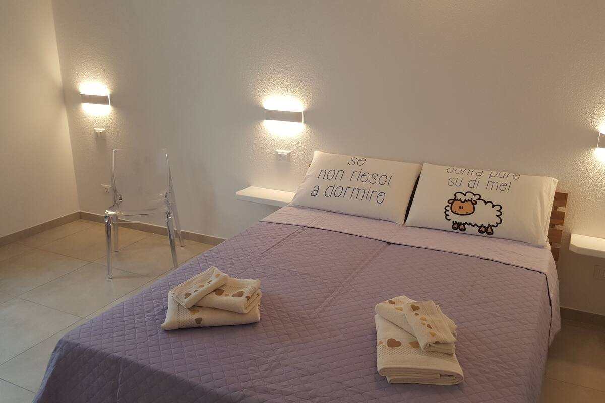 A neatly made bed with a purple quilt, two pillows with text and a sheep illustration, and two folded towels on the bed.