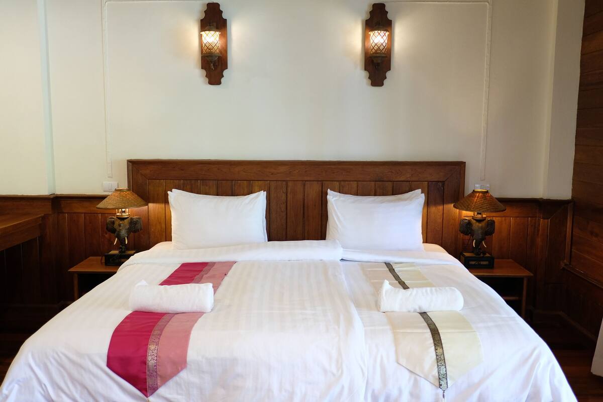 Un lit bien fait, avec du linge de lit blanc et des coussins décoratifs, deux lampes fixées au mur, une tête de lit et des tables de chevet en bois.