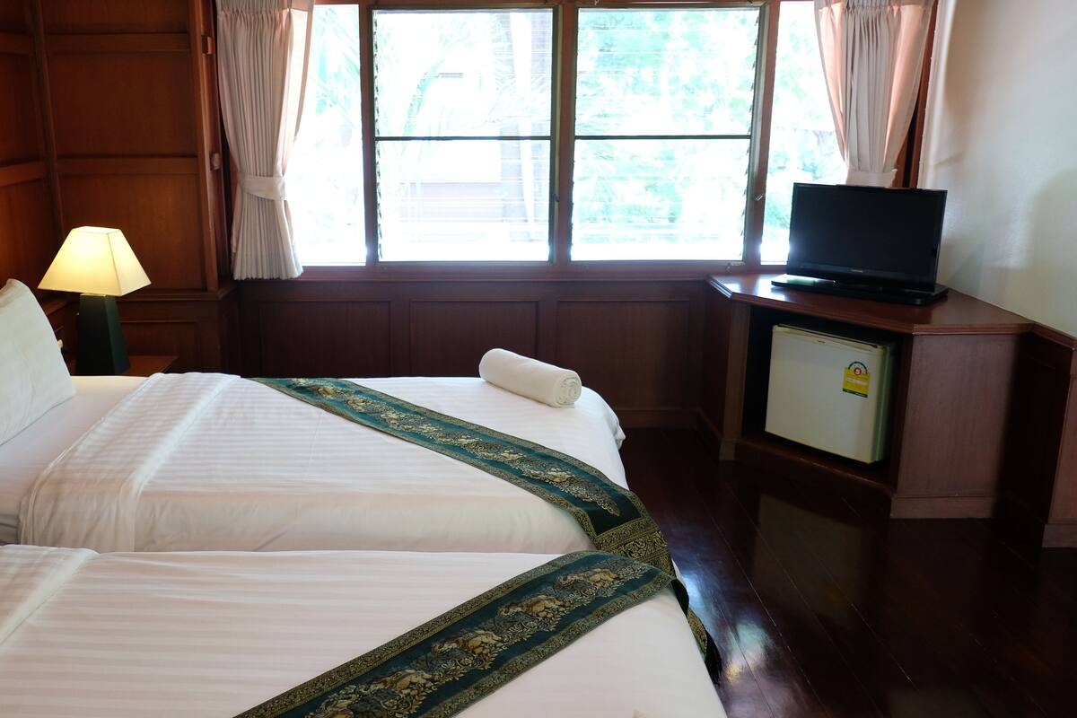 Une chambre d’hôtel avec deux lits, un téléviseur posé sur une commode en bois, une lampe et une fenêtre avec des rideaux.