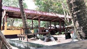 A covered pavilion with a tiled roof, wooden benches, and a sandy ground area with trees.