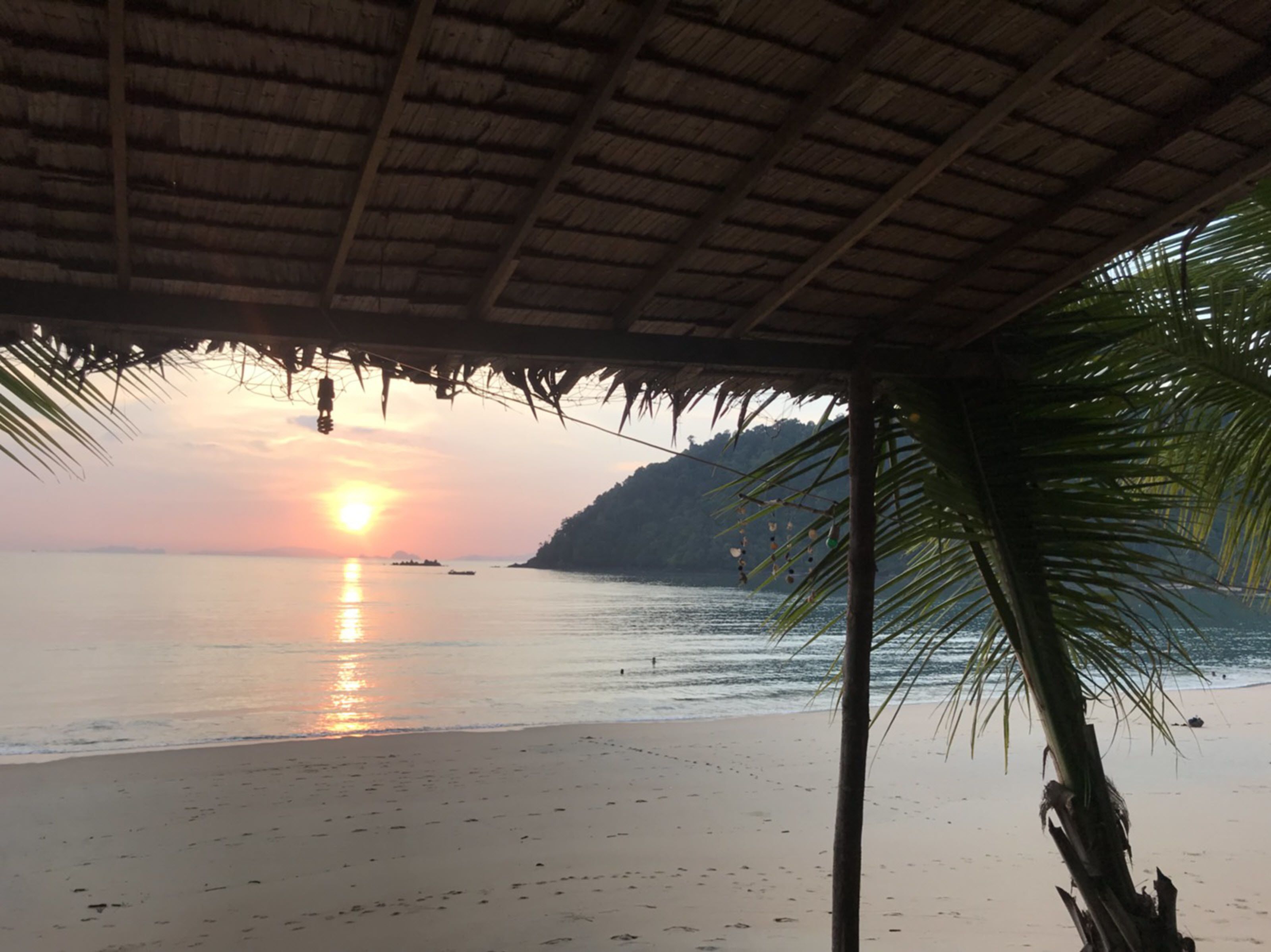 Une scène de plage avec une structure à toit de chaume, des palmiers et un coucher de soleil sur l’eau.