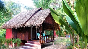 A rustic wooden house with a thatched roof, a porch with chairs, and a staircase leading to the entrance.