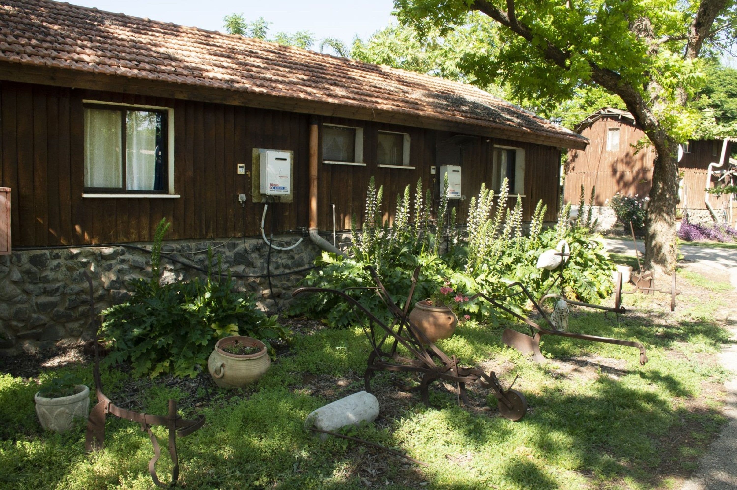 A rustic wooden building with a stone foundation, a tiled roof, and a garden area with potted plants and gardening tools.
