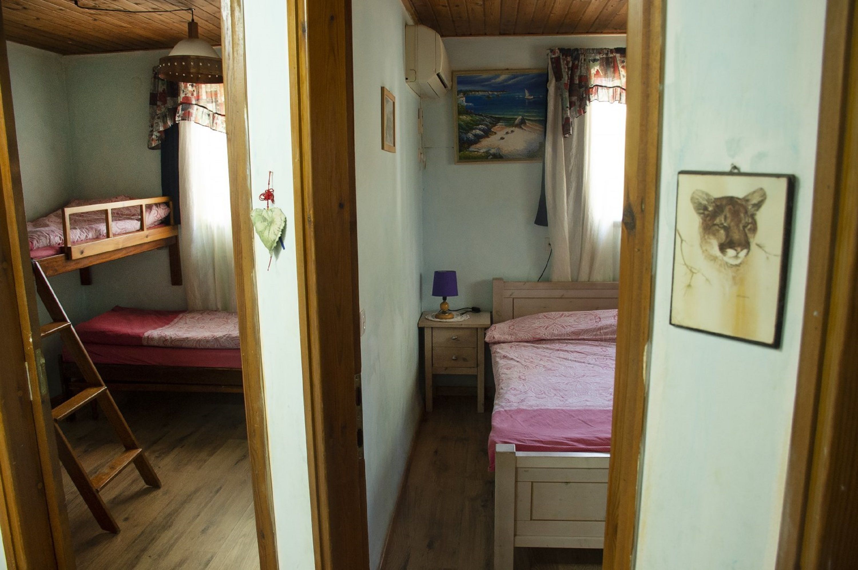 A narrow room with two bunk beds, a small bedside table with a lamp, and a painting of a beach scene on the wall.
