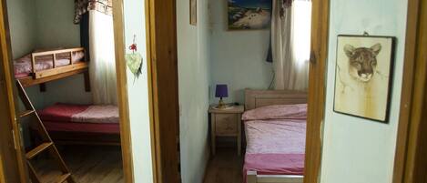 A narrow room with two bunk beds, a small bedside table with a lamp, and a painting of a beach scene on the wall.