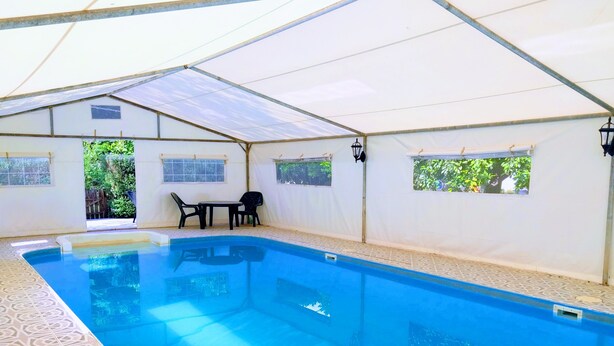 Une piscine intérieure avec un sol carrelé, un espace détente couvert comprenant deux chaises et une table, et des fenêtres donnant sur un paysage verdoyant.