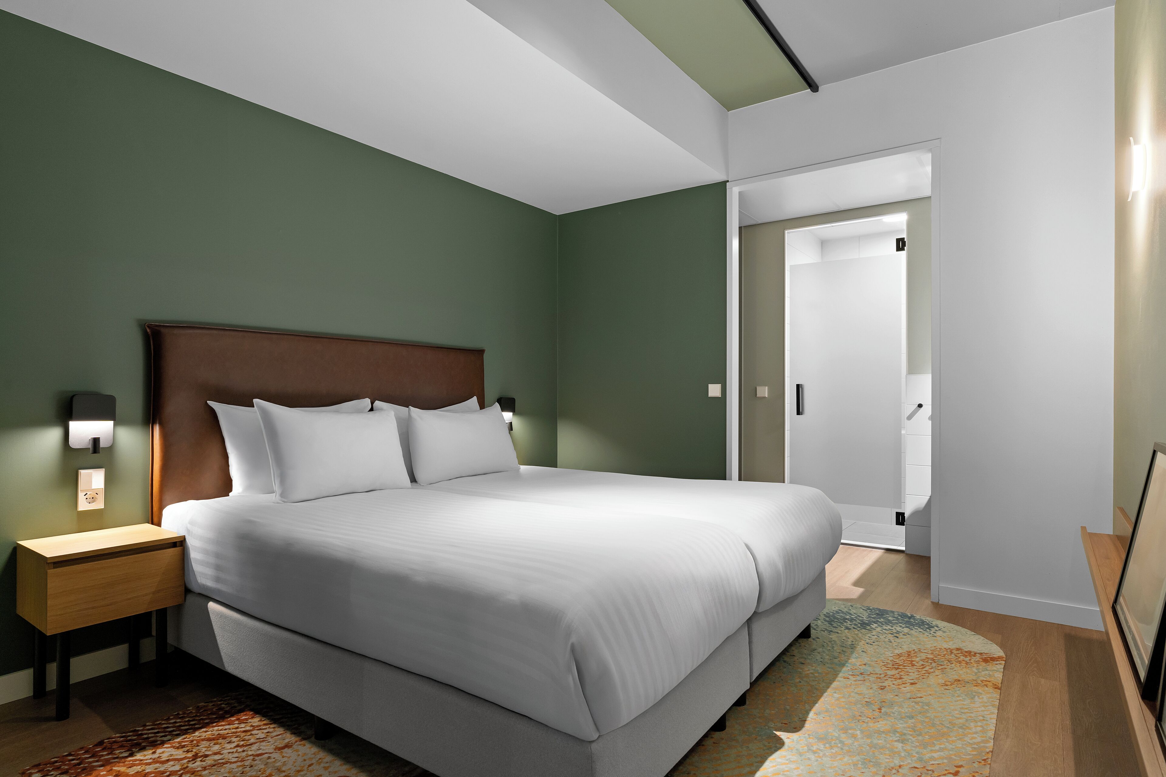 A modern hotel room with a large bed, a nightstand, a wall-mounted lamp, and a door leading to another room.
