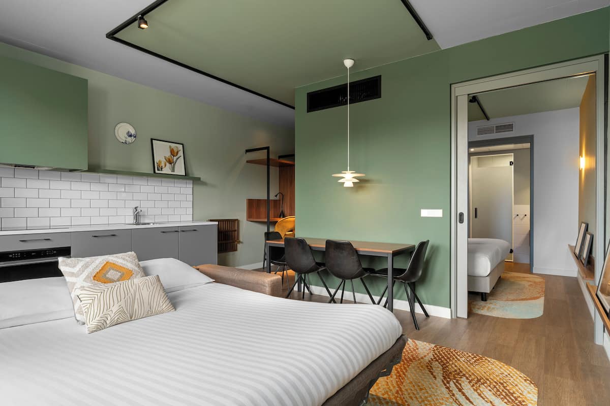 Ein modernes Hotelzimmer mit einem Bett, einem Schreibtisch und Stühlen, einer Küchenzeile und einem durch eine geöffnete Tür einsehbaren Badezimmer.