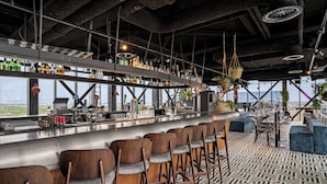 A bar area with wooden chairs, a bar counter with various bottles, and hanging plants. - Corendon Urban Amsterdam Schiphol Airport Hotel (Badhoevedorp)