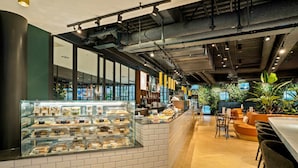 A modern cafe with a brick counter, seating area, and indoor plants. - Corendon Urban Amsterdam Schiphol Airport Hotel (Badhoevedorp)