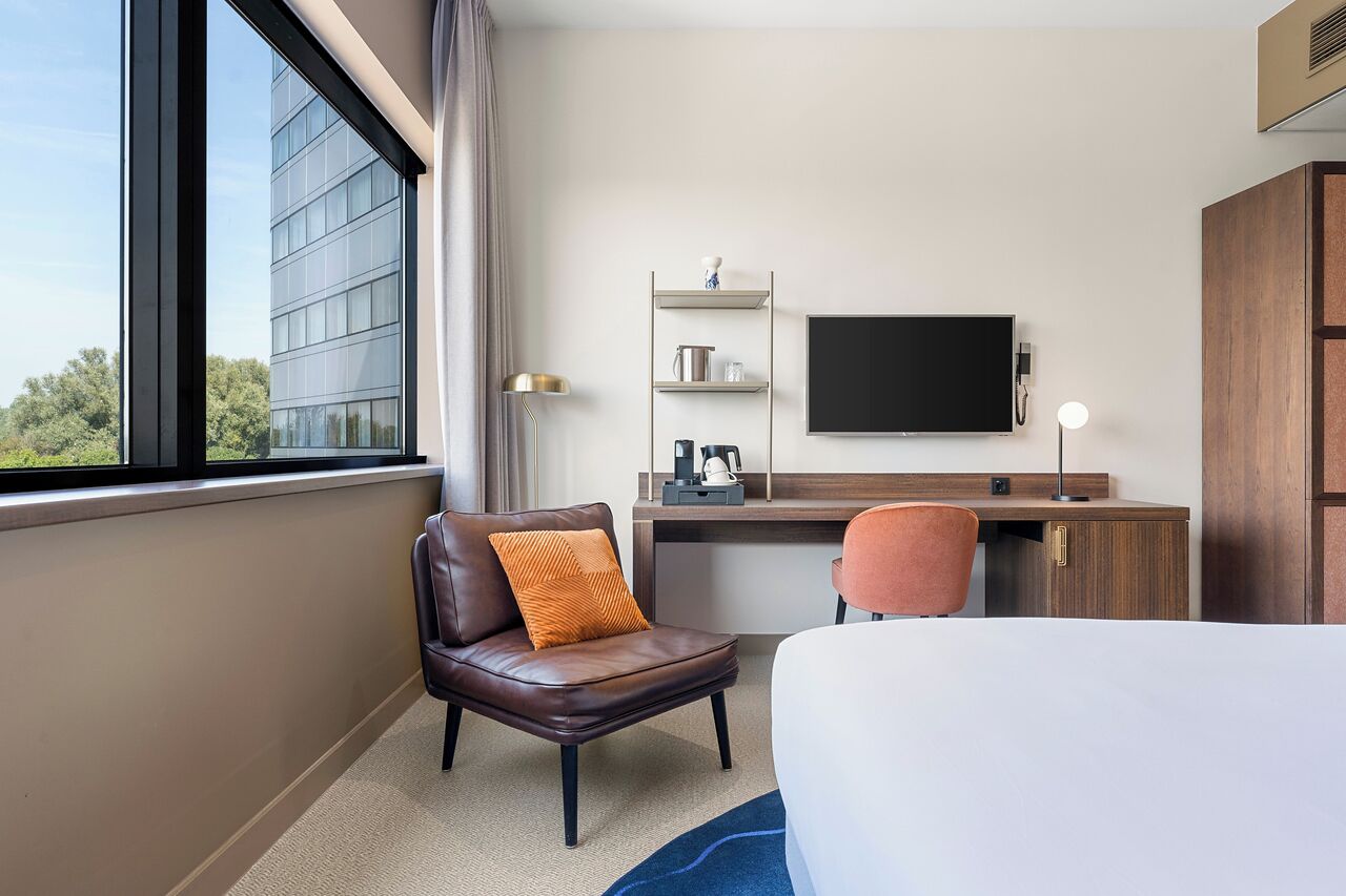 Habitación de hotel con cama, escritorio, silla, televisión y ventana con vista a un edificio.