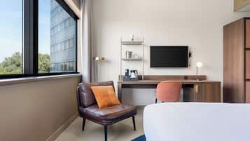 Habitación de hotel con cama, escritorio, silla, televisión y ventana con vista a un edificio.
