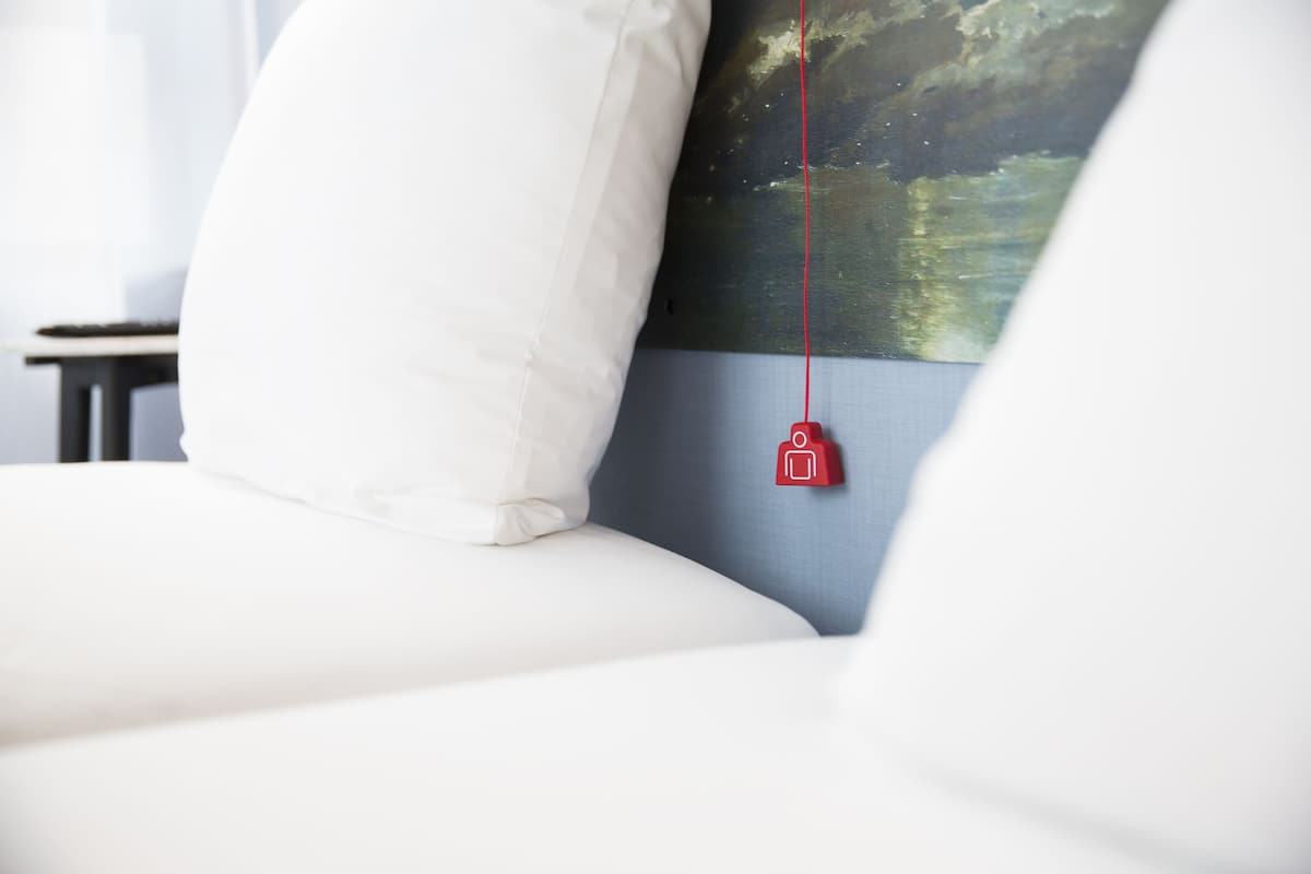 Ein Bett mit weißer Bettwäsche, ein rotes Objekt, das an der Kopfteil hängt, und ein Landschaftsbild an der Wand.
