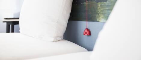 Una cama con ropa blanca, un objeto rojo colgando del cabecero y un cuadro de un paisaje en la pared.