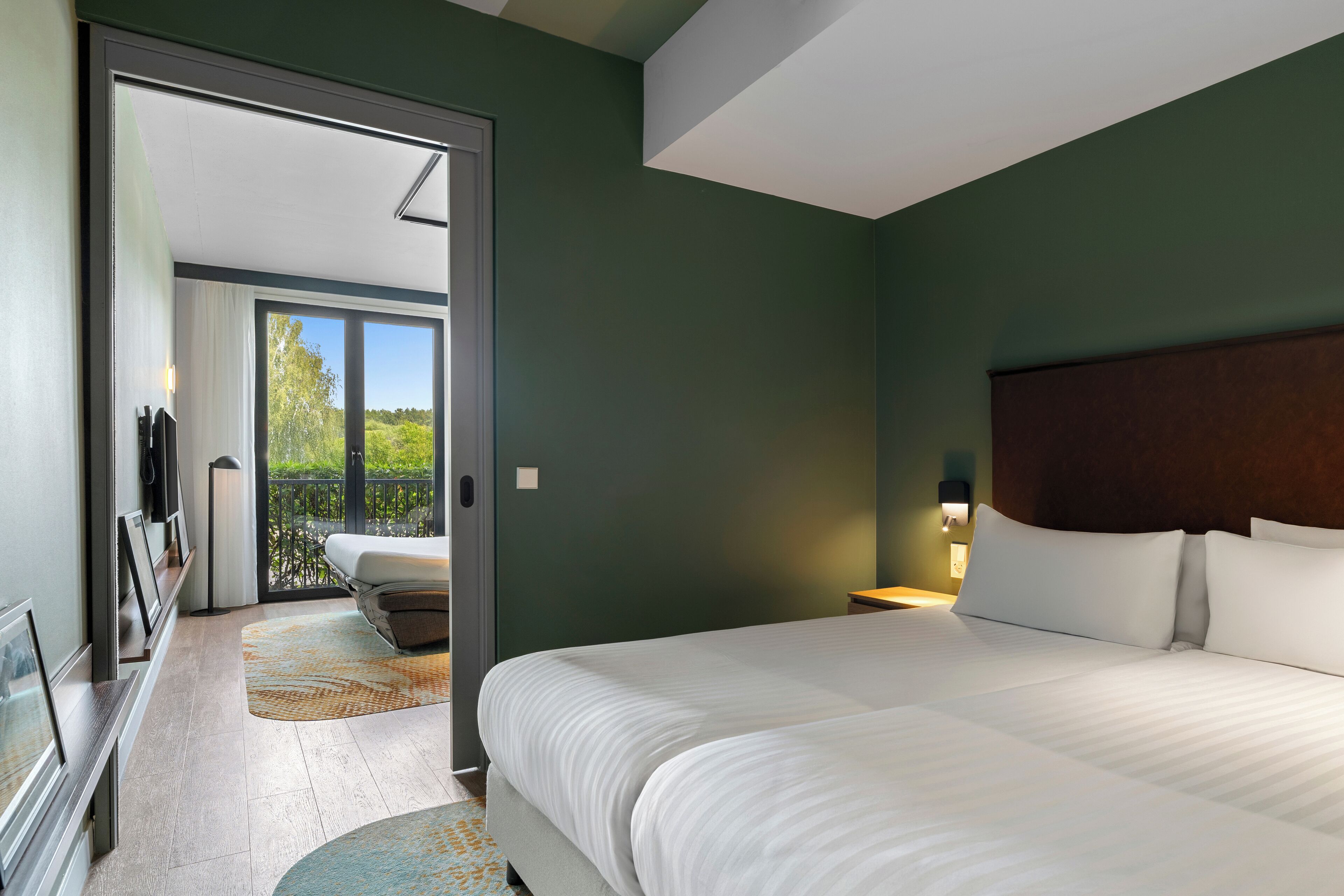 Un dormitorio con una cama grande, una mesita de noche con lámpara, una puerta que da a un balcón y vistas al verdor exterior.