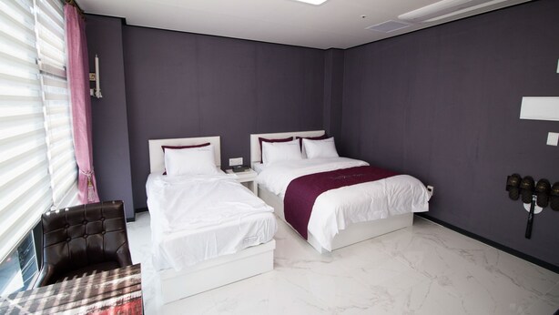 A hotel room with two beds, a nightstand, a chair, and a window with blinds. - The Stay Hostel (Yeosu)