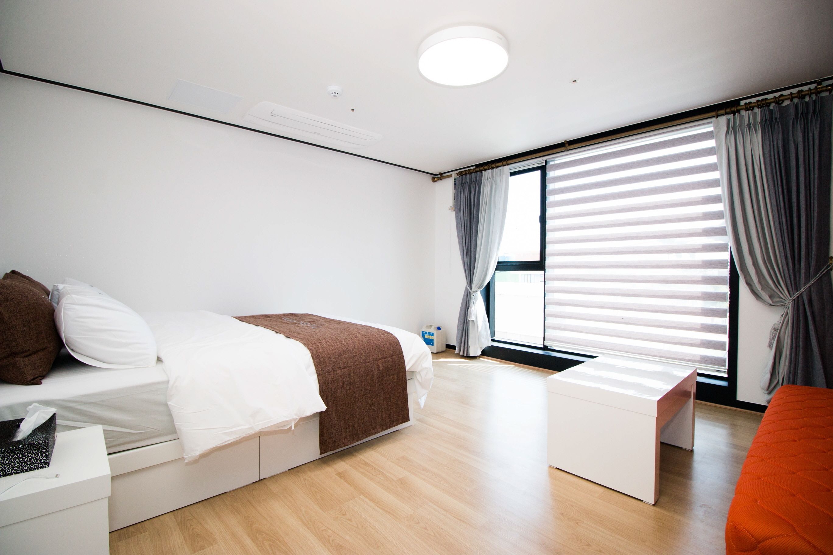Una habitación moderna y bien iluminada, con una cama, una mesita de noche y una pequeña mesita auxiliar blanca. Cuenta con una ventana con persianas y cortinas, y una lámpara de techo brillante.
