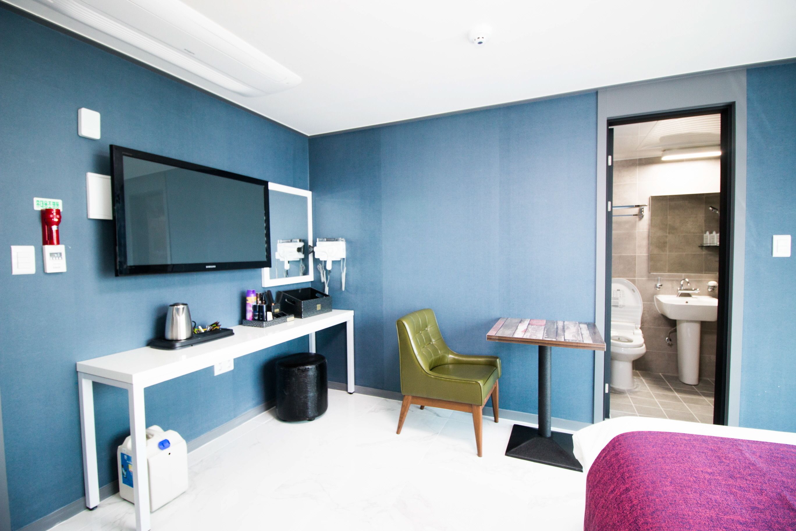 A hotel room with a bed, a desk with a flat-screen TV, a green chair, a small table, and a bathroom visible through an open door.