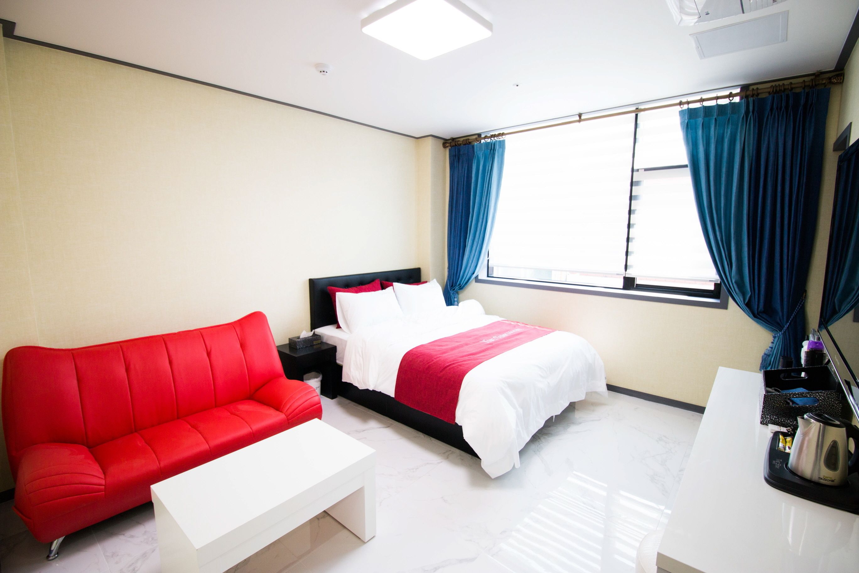 Quarto de hotel com sofá vermelho, cama com roupa de cama branca e vermelha, mesa de centro branca e escrivaninha com chaleira.