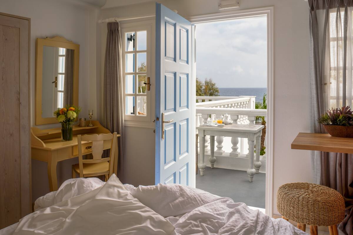 A bedroom with a bed, a desk, a chair, a mirror, and a view of the sea through an open door.