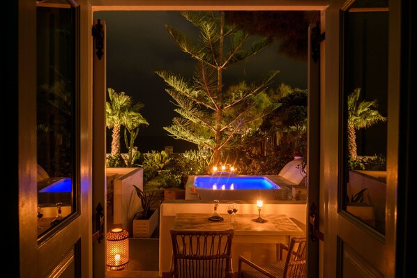 An outdoor dining area with a table, chairs, a candle, and a hot tub in the background. - Ocean Bay Suites (Santorini)