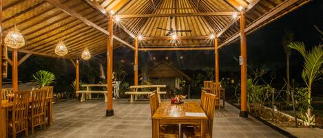An outdoor covered patio with wooden tables and chairs, a ceiling fan, and hanging lights.