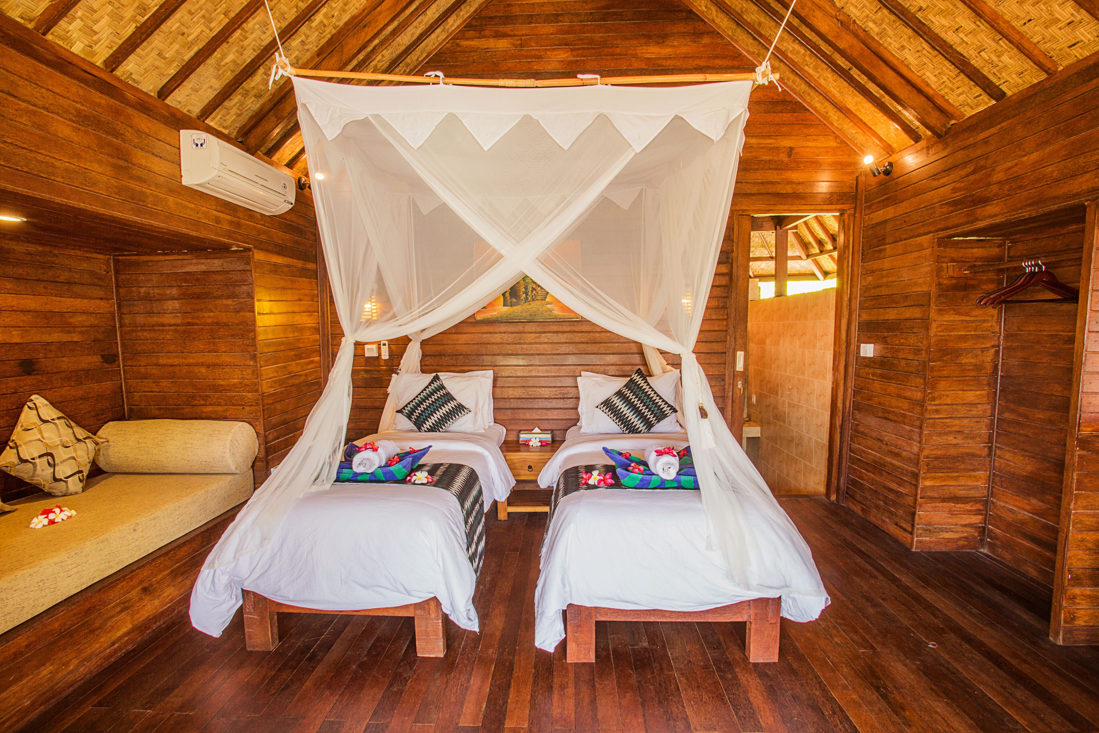A wooden cabin bedroom with two beds, a bench, and a hanging mosquito net.