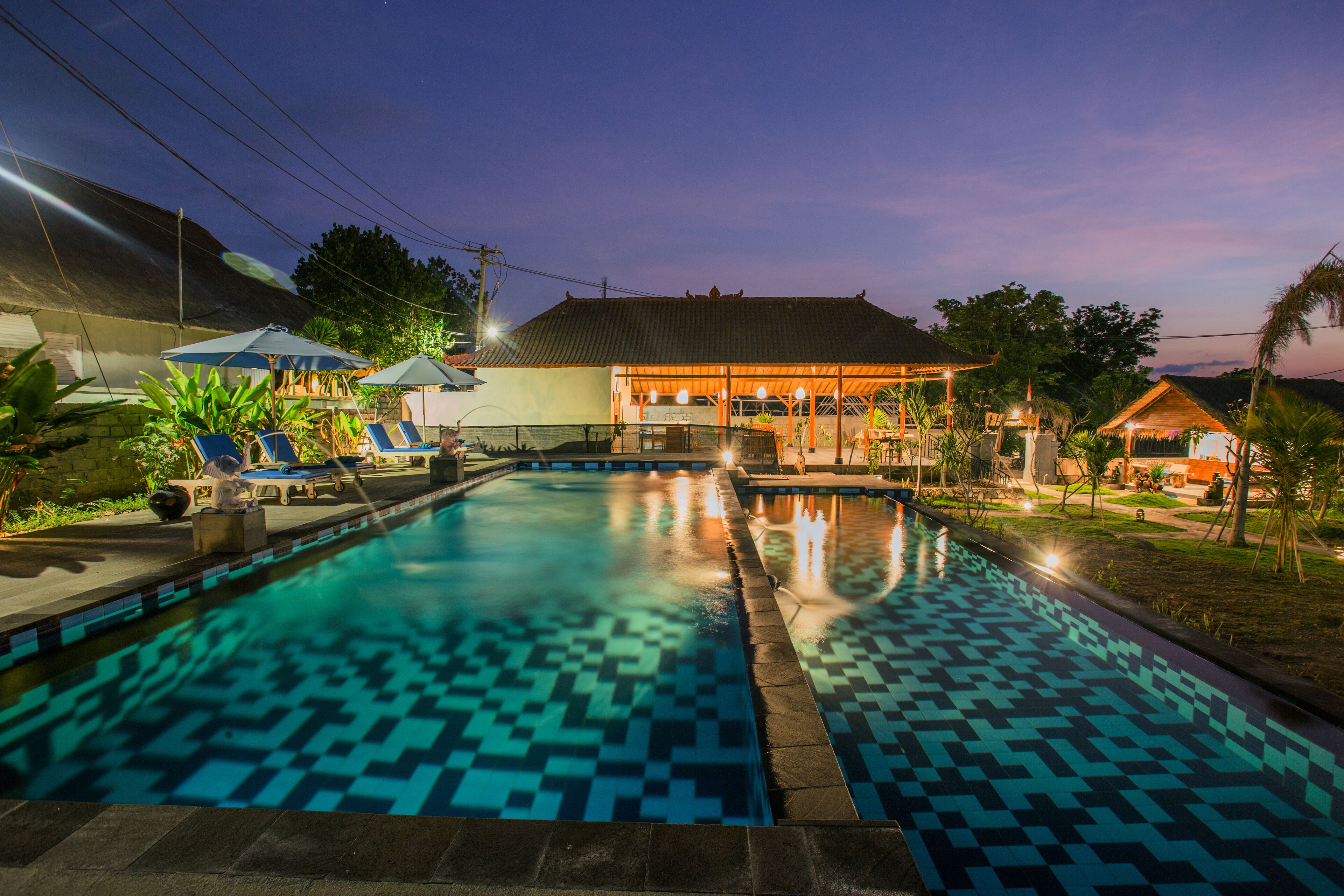 A swimming pool with a tiled pattern, surrounded by lounge chairs and umbrellas, with a building in the background.