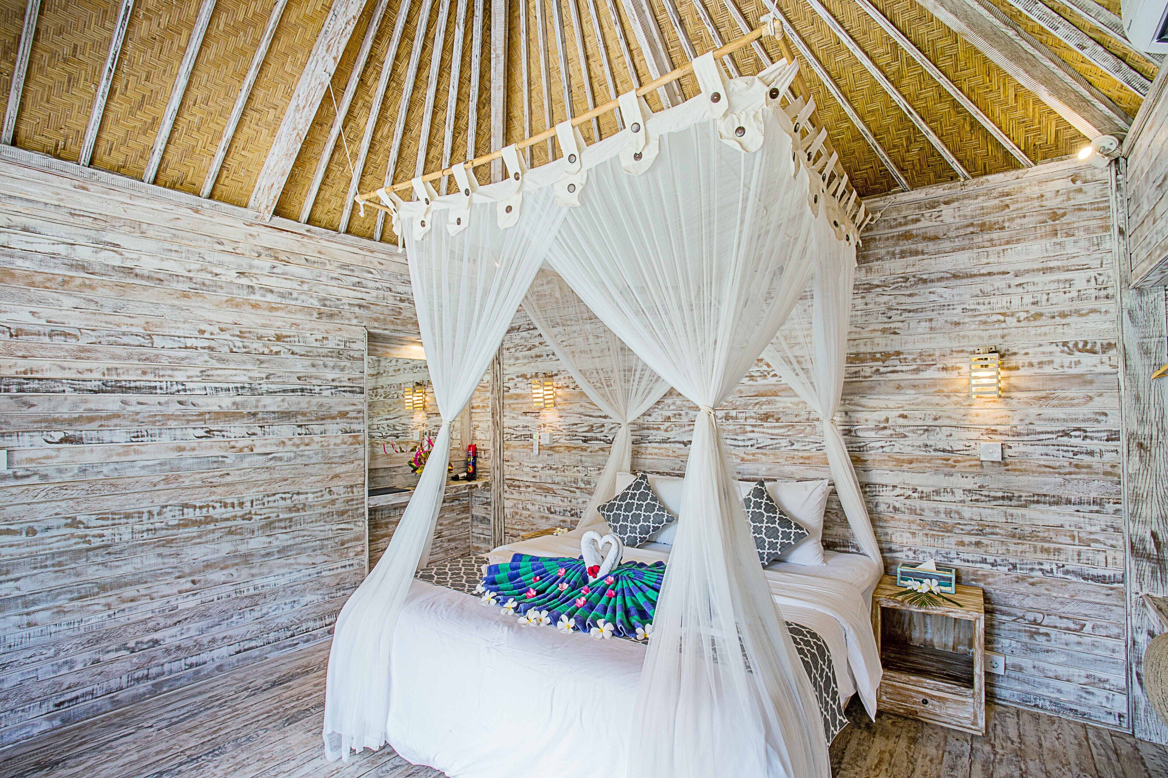 Una cama con dosel sobre cuatro pilares, con un dosel blanco, suelo de madera y pared de piedra.