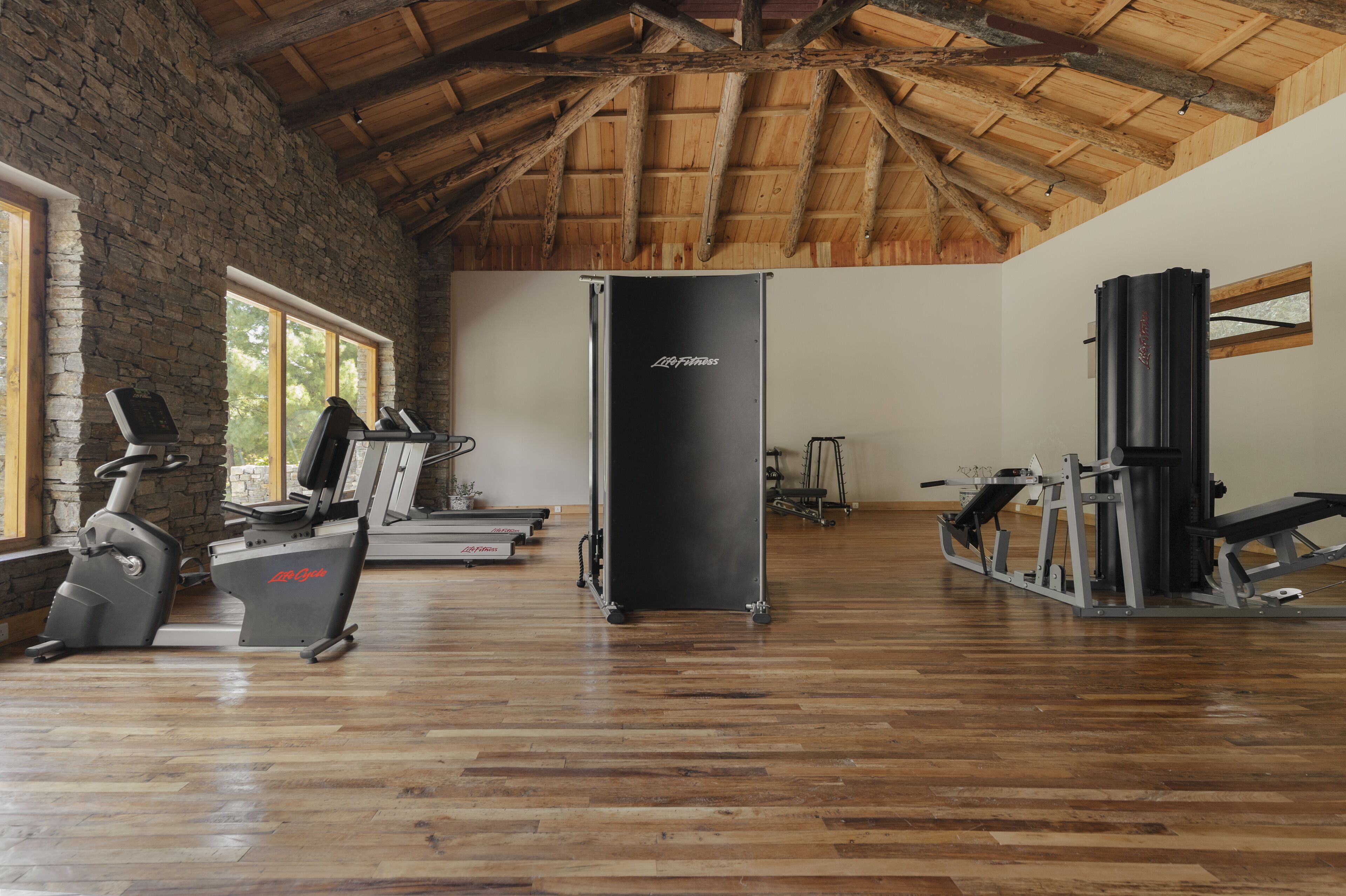 A gym with exercise equipment, a stone wall, wooden beams, and large windows.