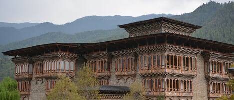 A traditional multi-story building with ornate wooden balconies and large windows, surrounded by lush greenery and mountains in the background.